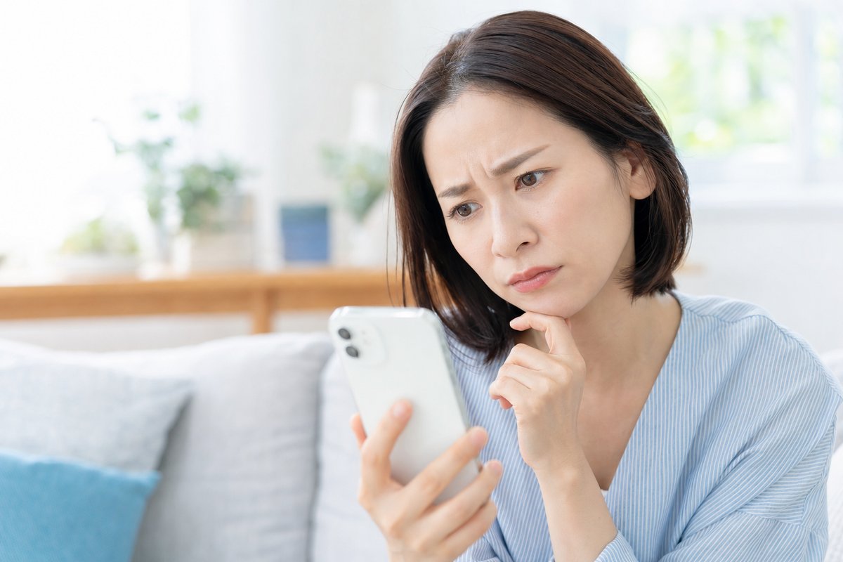 30代の女性が自宅のソファでスマートフォンを見ながら、眉を寄せて困った表情で考え込んでいる