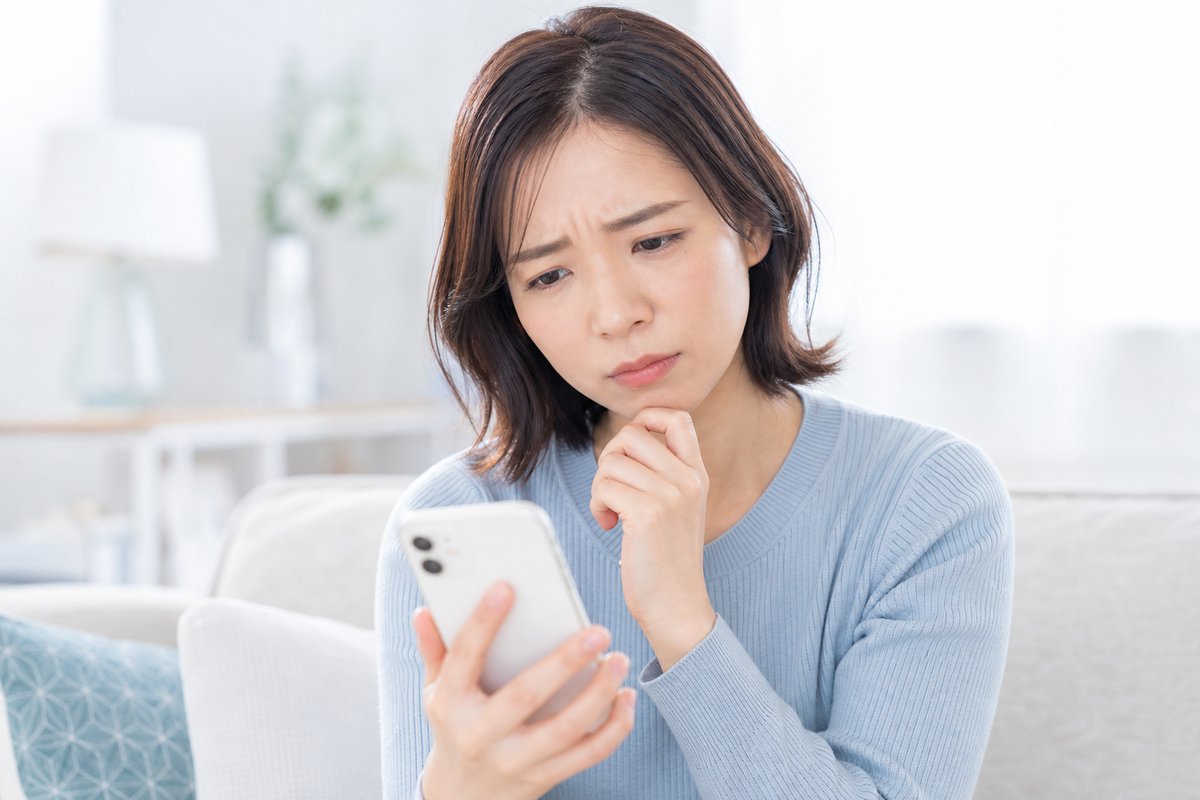 30代の女性が自宅のリビングでスマートフォンの画面を見ながら、眉を寄せて困った表情で考え込んでいる