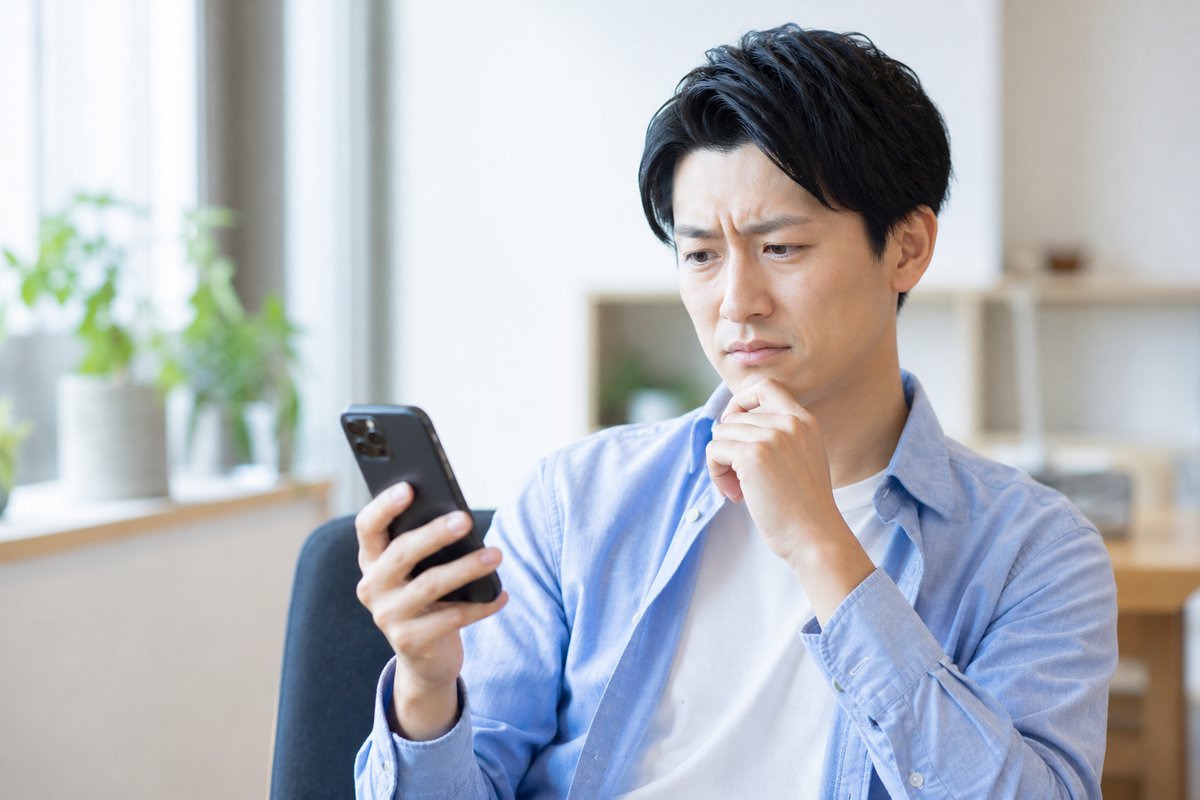 30代の男性が自宅の椅子に座り、スマートフォンを片手に眉をひそめて困った表情で考え込んでいる