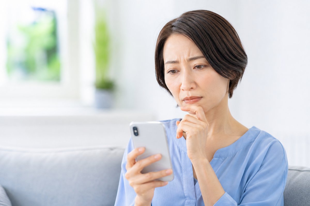 40代の女性が自宅のソファでスマートフォンを見ながら、眉を寄せて困った表情で考え込んでいる