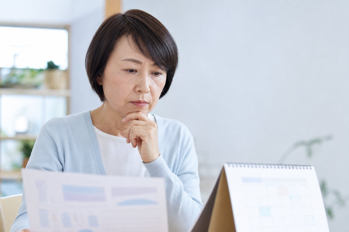 50代の女性が書類とカレンダーを前にして、不安そうな表情で顎に手を当てて考えている