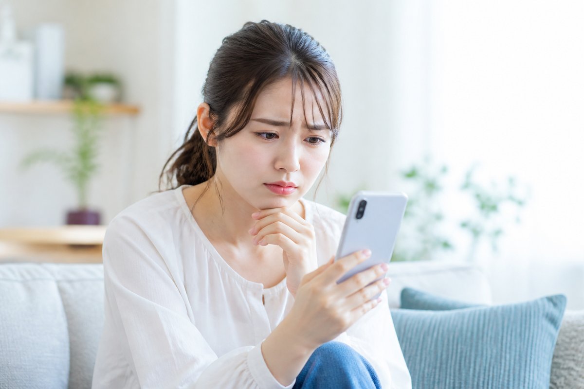 20代の女性が自宅のソファでスマートフォンの画面を見つめながら、眉をひそめて不安そうな表情を浮かべている