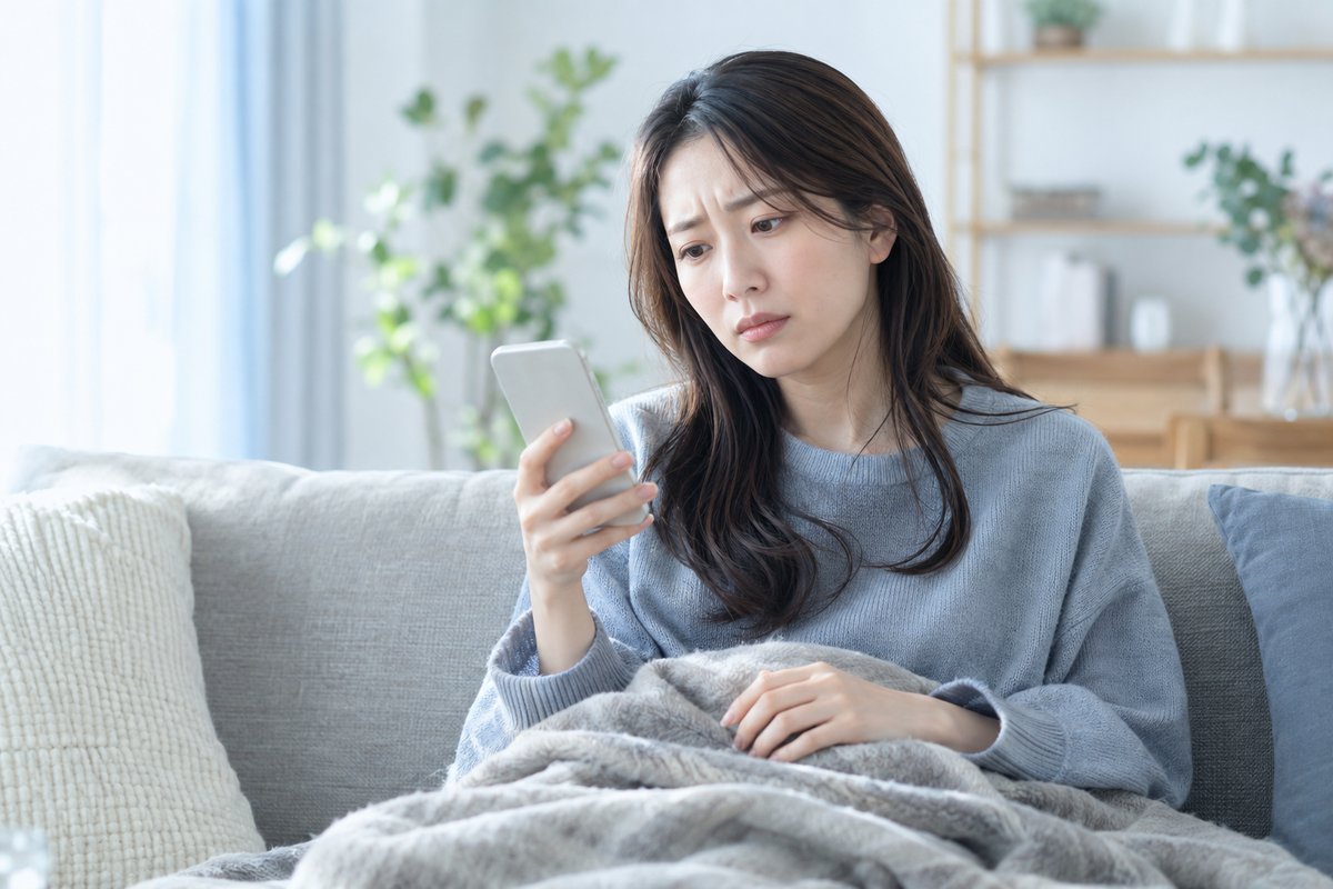 30代の女性が自宅のソファでブランケットを掛けたままスマートフォンを見つめ、眉を寄せて不安そうに考え込んでいる