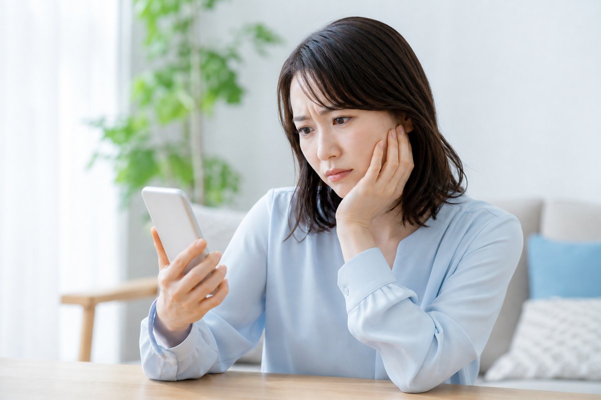 30代の女性が自宅のテーブルで、右手にスマートフォンを持ち左手を頬に当て、眉を寄せて考え込んでいる上半身のカット