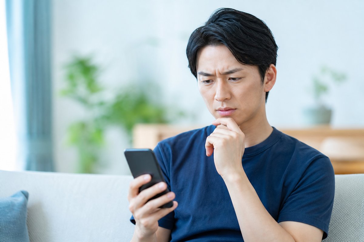30代の男性が自宅のソファでスマートフォンを片手に、眉根を寄せて困った表情で考え込んでいる